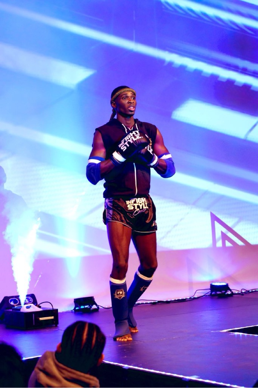 Robson Etuke Diamond walking out to the ring in a mongkol headband and InFight Style gear under blue and purple stage lights